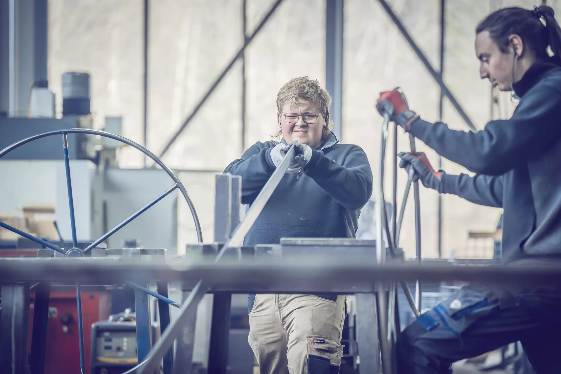 constructeur métallique fermant un oeil pour examiner une longue pièce de métal. Sur la droite une autre personne tient un cercle de métal ressemblant à un gouvernail