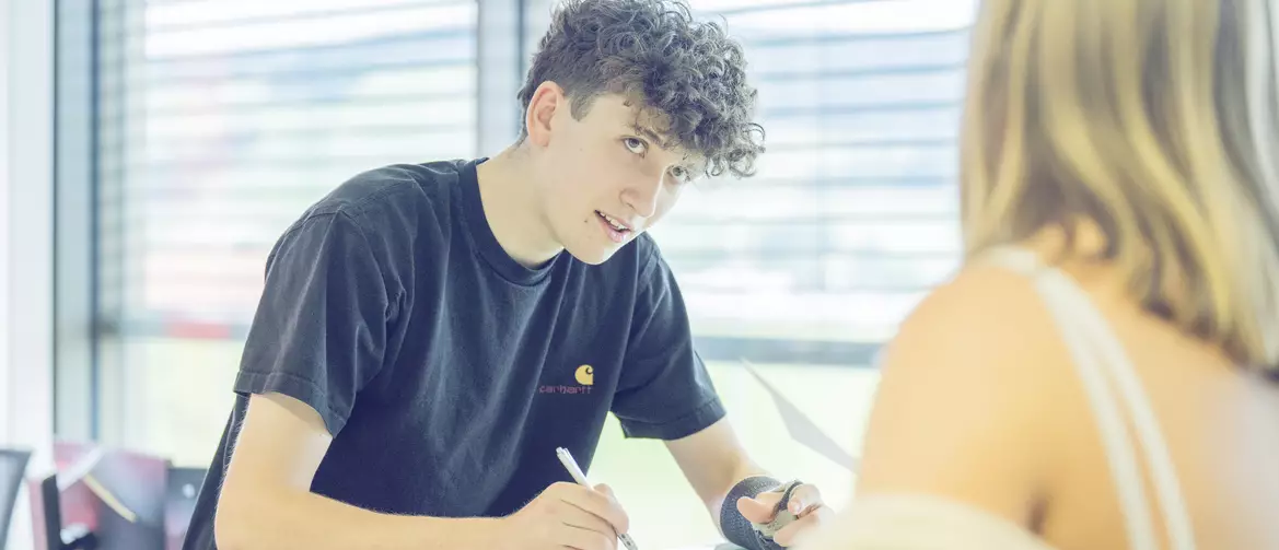 Un garçon regardant la fille qui est en face de lui et engageant la conversation. Il a une feuille sur laquelle il écrit avec un crayon papier.